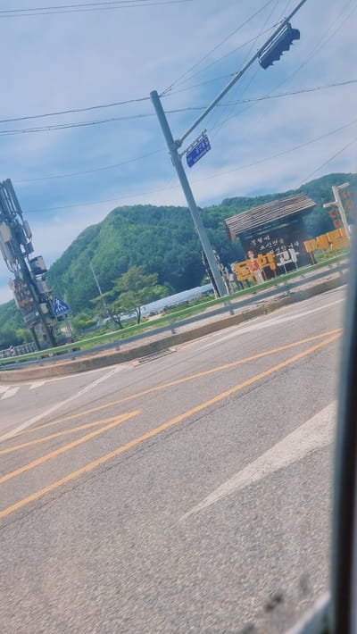 The image captures a view from inside a vehicle moving along a road in Yongpyeong-myeon, South Korea. The road has yellow lane markings. To the left of the road is a guardrail, beyond which lies a green mountainous area. There is also some building and infrastructure, including a traffic light and street signs written in Korean. The sky is visible above the mountain, with some cloud cover. A building is visible on the far left.