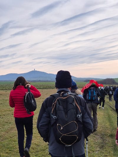 A group of individuals is observed walking along a grassy path in the vicinity of the city of Stara Reka, Bulgaria. The sky features extensive, linear altocumulus cloud formations. The immediate foreground features the back of an individual wearing a dark-colored padded jacket, a dark beanie, and a black hiking backpack equipped with light-colored elastic cord lacing. A single walking stick is visible extending downwards near their right side. To the left and slightly behind, a second individual, also observed from the rear, is dressed in a red puffer jacket and dark trousers, carrying a small black backpack by its strap over their left shoulder. A procession of additional individuals continues into the midground, all oriented in the same direction of travel. One subject within this group displays a blue backpack and a red textile object partially covering their head and upper back. The ground consists of a discernible path composed of packed earth and sparse grass, bordered by wider grassy fields. In the far distance, a prominent, green-tinged hill rises, topped by a tall, slender, cylindrical structure, characteristic of a communications tower.