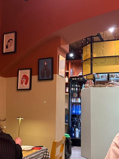 The image shows an indoor scene with an unusual perspective. A set of stairs and a wall adorned with framed pictures are visible on the left. A document rests on the stairs. Above, a decorated ceiling with lights is seen. To the right, a refrigerator stocked with blue-packaged goods and beverages stands, along with a table with a checkered cloth, and a person holding a thin golden object. The colors used are mainly brown, yellow, and white.
