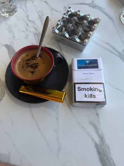 This is a top-down shot of a marble table setting that includes a pack of Parliament Aqua Blue cigarettes, an ashtray, a cup of coffee with a spoon, and a gold lighter. The cigarettes are in their original packaging, featuring the Parliament logo and "Smoking kills" in bold black text. The ashtray is metallic with multiple indented sections for cigarettes. The coffee is dark and frothy, served in a red-rimmed cup placed on a black saucer. The gold lighter is rectangular and rests on the saucer next to the coffee cup. The overall scene suggests a moment of relaxation or a break, likely indoors given the lighting. The text visible on the cigarette pack includes "PARLIAMENT", "Aqua Blue", "ORIGINAL RECESSED® FILTER", "ESTD 1931", "FIFTH AVENUE • NEW YORK", and "Smoking kills". The location context is Çanakkale Merkez, Turkey, which implies the setting could be a cafe, restaurant, or a home. There are no people visible in the image. The time of day and weather are not discernible from the visual information.