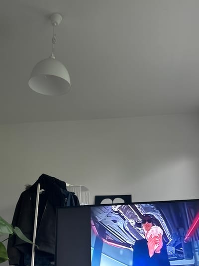 A white, dome-shaped lamp is mounted on a gray wall. To the right of the lamp is a television displaying a distorted image of people and objects with red and blue hues. A white shelf is attached to the wall beneath the television. On the shelf are several dark-colored garments and a plant with green leaves. The location in Gryfino, Poland is unknown.