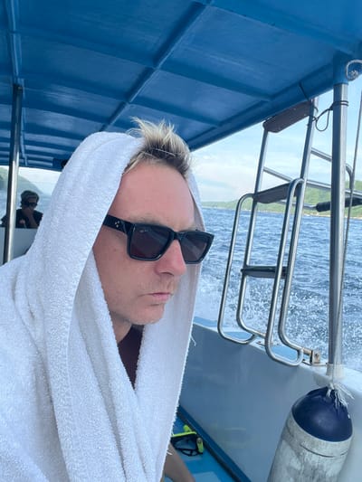 A man in sunglasses and a white towel rests on a blue boat. The vessel moves, evidenced by the water spraying behind its metal railing. A dark object hangs off the side. Another person sits in the background. The image is shot with the camera on its side.