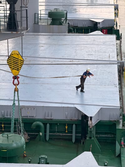 The image shows a worker on the deck of a ship, possibly in a port. The worker, wearing a white hard hat and dark overalls, is holding a red object and appears to be in motion. The deck is mostly metallic and appears to be a cargo ship given the presence of a crane hook with yellow and black stripes in the foreground. The background shows parts of the ship's structure and some industrial equipment. The overall scene suggests an active industrial or maritime environment. The prompt mentions Mersat El Hadjadj, Algeria, which would place this scene in a port facility within that region.