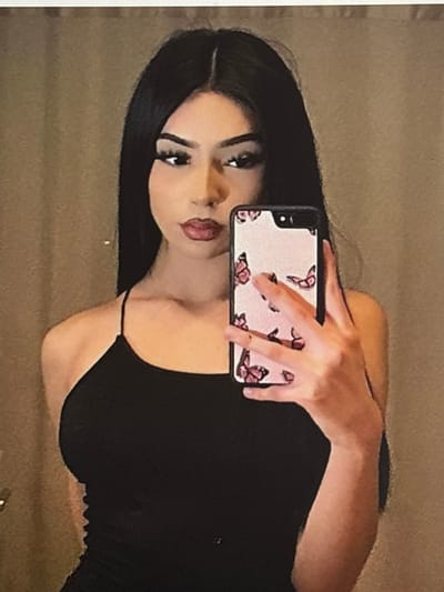 A woman with long dark hair poses in front of a mirror, holding a smartphone with a pink butterfly-patterned case. She wears a black top with thin straps. Her makeup includes defined eyebrows, eyeliner, and dark lipstick. She is positioned at an angle. Her expression is partially obscured by the phone. The background is a neutral, muted color.