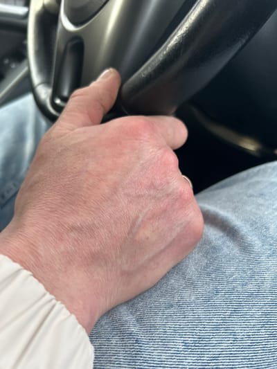 A close-up image depicts an adult's left hand resting on a vehicle's steering wheel, originating from the city of Ir'yak, Russia. The hand, exhibiting fair skin with visible veins and slightly reddened knuckles, is positioned with the thumb and index finger gripping the upper-right portion of a dark-colored, possibly black, steering wheel rim. A metallic ring is present on the ring finger. The steering wheel features a lighter, possibly silver-colored, inner spoke or trim. Below the hand, the individual is wearing light blue denim jeans. A portion of a light-colored, likely white or cream, long sleeve is visible on the left side of the frame, near the wrist area. The scene is illuminated from an unseen source, casting subtle shadows.