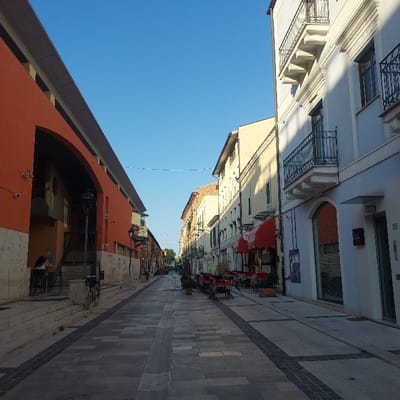 The image depicts a pedestrian street in Pescara, Italy, specifically in the Pescara Vecchia district, with the prominent orange Ex Aurum cultural center on the left. The scene is illuminated by clear daylight under a blue sky.

The street is paved with light-colored rectangular flagstones interspersed with darker patterned pavers. On the left, the multi-story orange building features a large, dark arched recess accessed via steps; two security cameras are visible on its exterior. Two individuals are seated at a table on a raised platform within this recess, engaged in an unidentifiable activity. Several bicycles are parked nearby the steps.

On the right, a row of multi-story buildings, primarily white, cream, and light blue, lines the street. Multiple ground-floor establishments display red retractable awnings over outdoor dining areas containing numerous empty tables and chairs. Upper floors feature balconies with black wrought-iron railings. The building on the immediate right is marked with the number '85' near its entrance. Black street lamps are visible at intervals. The street itself is largely devoid of pedestrian movement.