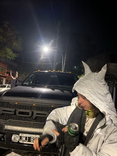 At night in Olivos, Argentina, a person wearing a white, pointed-hooded jacket is seen standing near a black Ford pickup truck. The truck has an Argentine license plate reading "JMJ 82". The person holds a green and black can. A street light is visible in the background.