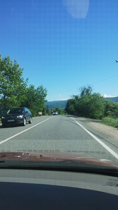 The image shows a road in Deliatyn, Ukraine, from the perspective of a vehicle. A black car is driving on the road, and another vehicle is visible further ahead. Trees line the side of the road. The sky is blue. Mountains are visible in the distance.