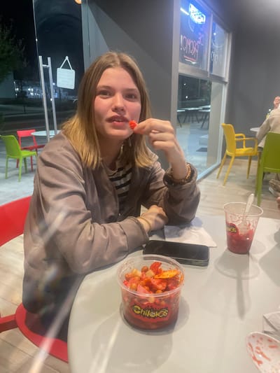 The scene depicts a young female inside an eatery in Fraccionamiento Los Heroes Queretaro, Mexico, during nighttime hours. She is seated at a light-colored table, facing the viewer, with shoulder-length light brown hair and wearing a grey jacket over a striped shirt. Her left hand holds a small, spherical red object to her mouth, while her right wrist displays a light-colored bracelet.
On the table in front of her, a clear plastic container labeled "Chilekos" is present, filled with a mix of red-seasoned peanuts, chips, and other snack items. To its right, a black smartphone rests on a crumpled white napkin. Further to the right, a clear plastic cup containing red residue and a white plastic spoon is visible.
The background features large glass windows revealing a dark exterior, indicating night. Through these windows, distant streetlights, an outdoor seating area with red and bright green chairs, and a white hanging "open/closed" sign are discernible. Inside the establishment, partial views of other patrons and brightly colored yellow and lime green chairs are visible, along with neon signs, one stating "PROMO!," illuminated through the windows.