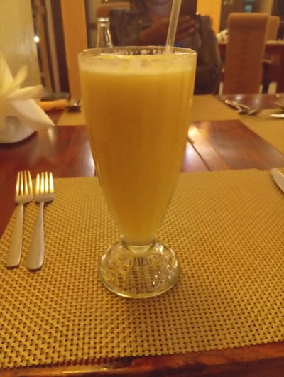 This image captures a close-up of a dining table, likely within a restaurant or cafe during the evening. The central subject is a tall, clear glass filled with a yellow-orange beverage, possibly a smoothie or juice, with a straw inserted. A light layer of foam or froth is visible at the top of the drink.
The glass is placed on a light brown, rectangular woven placemat, which covers a dark, polished wooden table. To the left of the glass, two silver forks are neatly arranged side-by-side on the placemat. Further left, a white napkin is elegantly folded in a fan shape and placed on a white plate, indicating a complete place setting.
The background is softly blurred, revealing an indoor setting with warm, artificial lighting, suggesting it's either nighttime or a dimly lit interior. A person is faintly visible in the upper background, appearing to be seated and possibly holding a phone. Other blurred elements include parts of wooden furniture, such as chairs and additional tables, contributing to the ambiance of a dining establishment. There are no discernible location cues within the image itself beyond the general restaurant setting, but the context indicates it was taken in Makuyu, Kenya. No text is visible in the image.