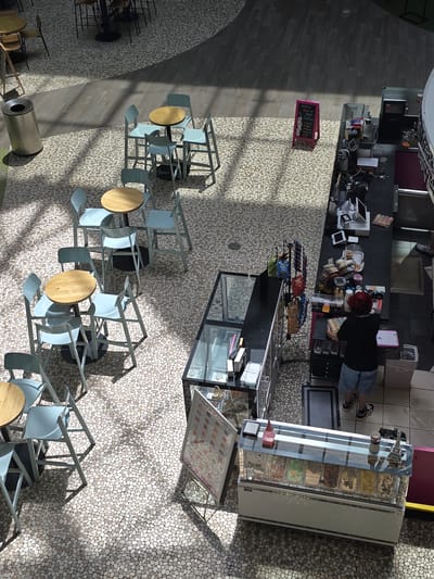 The image shows an aerial view of an outdoor seating area adjacent to a food service establishment, likely a cafe or ice cream parlor, in Alpharetta, United States of America. The ground is paved with small, rounded pebbles. Several small, round tables with light-colored wooden tops and light blue chairs are arranged in clusters. A silver cylindrical trash receptacle is visible to the left. Further to the right, a counter area is set up with a person wearing a black t-shirt and denim shorts behind it, handling a package. In front of the counter, a glass display case showcases various tubs of ice cream. A pink chalkboard sign listing coffee drinks stands near the seating area. Sunlight casts distinct shadows across the scene.