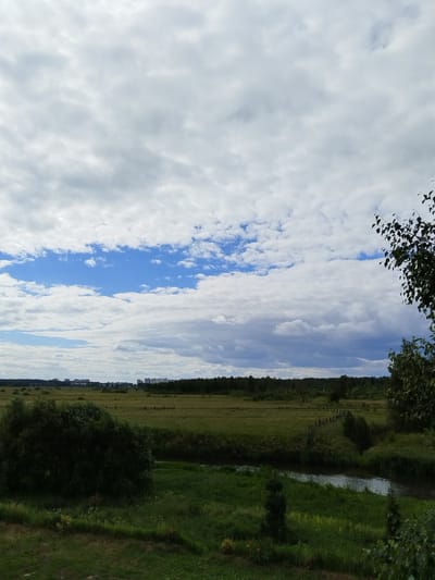 The image depicts a wide, panoramic view of a rural landscape, likely on the outskirts of Sverdlovskiy, Russia. The foreground features lush green grass and foliage, with a small, winding stream or river to the right. A large, dark green bush is prominent in the left-center of the frame. In the mid-ground, a vast, green field stretches towards a line of dark green trees that form the horizon. Faint, wooden posts are visible in the field, suggesting a fence or boundary. In the distance, a cluster of modern, multi-story buildings can be seen, indicating the presence of a town or city. The sky above is a dramatic canvas of clouds, with patches of bright blue sky peeking through. The overall impression is one of a serene, natural setting juxtaposed with the signs of urban development. There are no people or animals visible in the image.