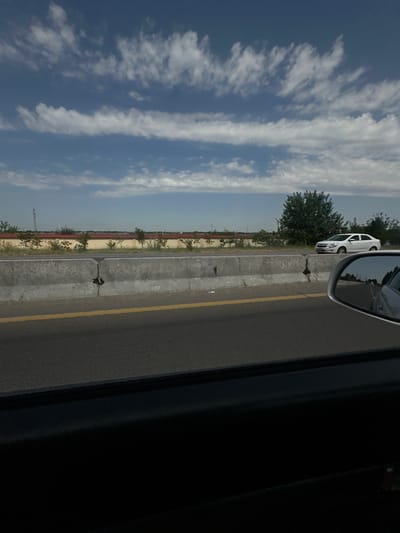 The image depicts a view from inside a vehicle traveling on a highway. The foreground is dominated by the interior of the car, with a dark, reflective surface on the bottom and the side mirror on the right. The highway itself occupies the mid-ground, with a concrete barrier separating the lanes of traffic. A single yellow line marks the edge of the lane. Beyond the barrier, a landscape stretches out, featuring a row of trees and shrubs, a yellow wall, and distant structures that appear to be industrial, possibly power lines or towers. A single white car is visible on the highway, traveling in the same direction as the camera. The sky above is a blend of blue and white, with scattered clouds. The location is specified as the city of Istiqlol, Uzbekistan.