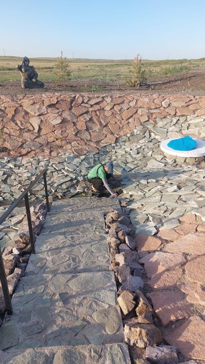In Zhartas, Kazakhstan, a person wearing a green shirt and dark pants kneels on a stone pathway, appearing to tend to something on the ground. A plastic water bottle rests nearby. The pathway leads down from a rocky embankment constructed of large, irregularly shaped stones. Above the embankment, a grassy field stretches towards the horizon under a clear blue sky. To the left, a seated statue is visible, and in the distance, sparse vegetation dots the landscape. A white circular platform with a bright blue object on top is situated on the rocky embankment.
