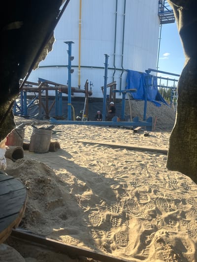 The image depicts an outdoor industrial construction site. In the foreground, sand covers the ground, marked with numerous footprints. Several buckets, one metal and one red plastic, are present, along with some cylindrical objects that appear to be concrete pipes or rolls of material. In the mid-ground, there is a blue metal structure supporting various pipes and equipment. Two individuals are visible in this area; one wearing a hard hat and dark clothing, possibly engaged in work, and another sitting, wearing a white cap and dark clothing. In the background, a large, white, cylindrical industrial tank dominates the scene, with a yellow pipe running vertically alongside it. Blue tarps are draped over a pile of gravel. The overall environment suggests ongoing construction or maintenance work at an industrial facility. The location is identified as Chikshino, Russia.