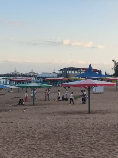 This is an eye-level, medium shot of a sandy beach scene in Bayet, Kyrgyzstan. In the foreground, a sandy beach is populated by several people and numerous colorful beach umbrellas, some of which are open. A few people are gathered under a large red and white umbrella in the center, and others are scattered across the sand, some sitting, others standing. There are at least six visible beach umbrellas in various colors, including red and white, green, blue, and yellow. In the middle ground, a body of water, possibly Lake Issyk-Kul, is visible, with a pier extending into it. Several structures with domed roofs and other beach-related buildings are situated along the waterfront. The sky above is a pale blue with scattered, wispy clouds. Two birds are present in the sky, one further in the distance than the other. The overall atmosphere is that of a recreational beach on a clear day.