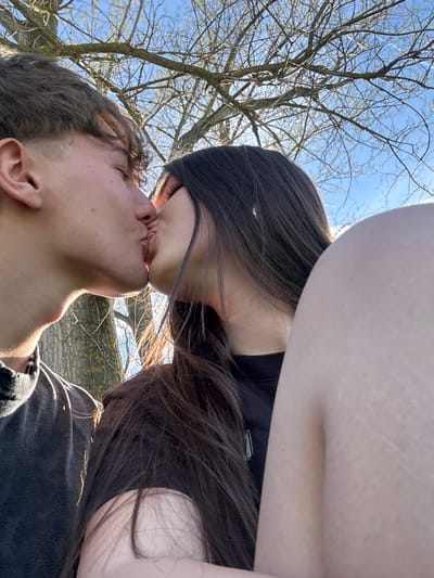 A low-angle close-up shot shows two people kissing outdoors. One person, with short brown hair and fair skin, is lying down, facing upward. The other person, with long dark hair, is leaning down to kiss them. Bare tree branches and a blue sky are visible in the background. The location of Beli Brod, Bulgaria, cannot be confirmed.