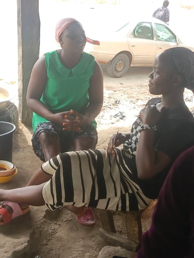 Two women are conversing outdoors. One woman, wearing a green top, sits on the ground. The other, in a black shirt and striped skirt, sits on a wooden stool. A car is visible in the background, with a person walking behind it. Several bowls are near the first woman. The setting appears to be a casual gathering in a rural or semi-rural area.