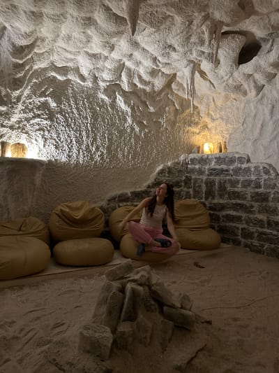 In a salt cave, a young woman sits cross-legged on the floor, her head tilted back and eyes closed, with a relaxed expression. She is wearing a white tank top and pink pants. The cave walls are rough and textured, with formations resembling stalactites hanging from the ceiling. Soft lighting emanates from behind stone structures and in cylindrical lamps on shelves, casting a warm glow. Beanbag chairs are arranged in a semi-circle behind her. The floor is covered in what appears to be salt crystals, with a small pile of salt rocks in the foreground. The environment suggests a therapeutic or relaxation space. The location is identified as a salt cave in Korsuntsi, Ukraine.