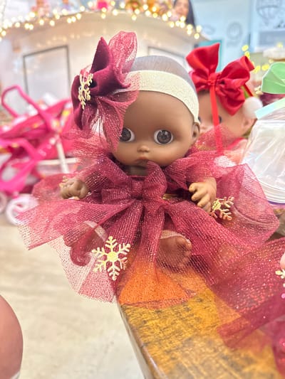 A dark-skinned doll with large dark eyes and a small mouth is depicted seated on a polished wooden surface in an indoor commercial setting within Santa Cruz Aquiahuac, Mexico. The doll is dressed in a sleeveless, tiered garment made of burgundy glitter tulle, featuring multiple matching bows and gold snowflake appliqués. A broad, light-colored headband with a large burgundy bow and a gold snowflake ornament is positioned on its head. Its small hands are visible near its torso, and its legs are bent, revealing small feet. In the immediate background to the left, blurred pink toy strollers are visible. To the right, a second doll with a red head bow is partially visible, alongside clear plastic packaging. The distant background consists of a white display structure, blurred figures, and warm-toned string lights, indicative of a toy store or market stall.