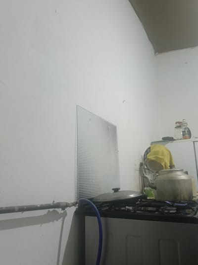 The image depicts a corner of what appears to be a kitchen. A white wall occupies the majority of the frame, with a gray ceiling visible in the upper right. On the wall, there is a glass panel with a dotted pattern. To the right, a gas stove is visible with a pot on it, and a kettle with a yellow cloth draped over it. There are also some bottles on a shelf in the background. A gas pipe runs horizontally across the left side of the image, with a blue hose attached. The scene suggests a domestic setting, possibly in a modest kitchen. There are no people visible in the image. The overall impression is of a functional, albeit somewhat cluttered, kitchen space. Without further context, it is impossible to definitively place this scene within the city of Altinkul, Uzbekistan.