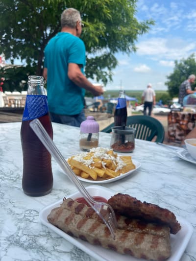 The image depicts an outdoor dining scene in Hitrino, Bulgaria. In the foreground, a white marble-patterned table holds a clear glass bottle of dark soda, 250ml in volume, with a blue label featuring Cyrillic script and condensation on its surface, containing a transparent plastic straw. Beside it is a white rectangular foam plate with two grilled oblong meat items, one pierced by a transparent plastic fork, and a small amount of red condiment (ketchup) visible. Adjacent is a white round plate containing french fries topped with crumbled white cheese. To the left, a small, light purple lidded container holds a translucent liquid. To the right, a small glass jar contains red flakes, and a portion of a dark green slatted plastic chair is visible. In the midground, out of focus, a man in a teal t-shirt and glasses stands facing away from the camera, beneath green foliage. The background features a bright sky with scattered white clouds, more blurred figures, trees, and distant structures including a white vehicle and tents, suggesting an open public area.