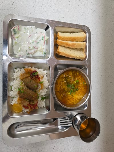 A metal cafeteria tray holds a meal consisting of four compartments. The top left compartment contains three slices of white bread. The top right compartment holds a bowl of soup with noodles, meat, and herbs. Next to the soup, a metal mug is filled with a light-colored liquid, likely tea. Below the soup and mug, a fork and spoon rest in a long, shallow compartment. The bottom left compartment contains a creamy salad with chopped radishes and herbs. The bottom right compartment features a mound of white rice topped with two meat patties and a mixture of vegetables, including onions and tomatoes, garnished with fresh herbs. The tray is placed on a speckled countertop. The overall scene suggests a meal served in a cafeteria or dining hall setting, possibly in Kyrgyzstan or Uzbekistan, given the provided location context. The lighting is bright, indicating daytime. No people are visible in the image, and the focus is solely on the food and the tray. The metal tray has a visible brand name, "KAILAKI," stamped on its surface.