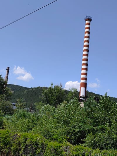 The image shows two industrial chimneys against a backdrop of a blue sky with scattered clouds and a densely forested hill. The chimney on the right is tall and painted with alternating red and white stripes. It has a metal railing at the top. The chimney on the left is shorter, appears older, and is partially obscured by trees. The foreground is filled with lush green vegetation, including trees and bushes, with a hint of a fence at the bottom. The scene suggests an industrial site located in or near the city of Belovo, Bulgaria, given the presence of industrial chimneys.