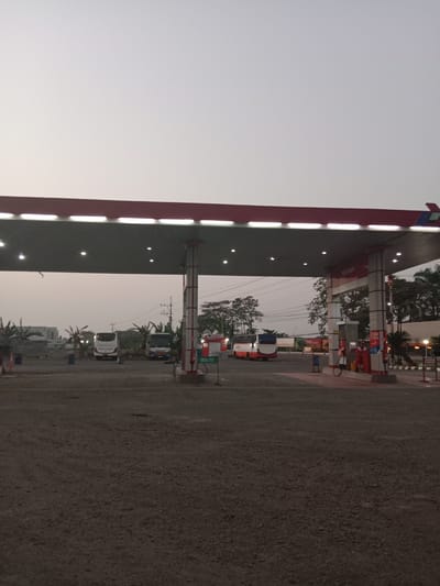 This image captures a gas station in Brangsong, Indonesia, under a pale, overcast sky that suggests either dawn or dusk. The foreground is a rough, gravelly surface, characteristic of many industrial or commercial outdoor areas. Dominating the mid-ground is a red and gray gas station canopy, brightly lit by overhead fluorescent lights.

Under the canopy, a red and white gas pump is visible, with a hose and nozzle attached. To the right of the pump, a white pole with red and white stripes supports the canopy. A green sign with white lettering that reads "DEXLITE" is positioned near the pump.

In the background, several vehicles are present. A white bus with tinted windows is parked in front of a low-rise building with a dark roof. To its right, a blue and white bus is also parked, and further back, a large truck is visible. Palm trees and other vegetation are scattered around the area, adding a tropical feel to the scene. The overall impression is one of a functional, outdoor service area, likely a busy transit point given the presence of multiple buses and trucks.