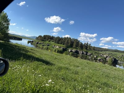 The image depicts a scenic rural landscape on a sunny day, featuring a body of water winding through rolling green hills. The foreground is a field of lush green grass dotted with white wildflowers, with a dark car side mirror visible on the left edge. The middle ground showcases the water, bordered by a rocky shoreline covered in trees and shrubs. The background reveals more hills and a clear blue sky with scattered white clouds. The overall impression is one of natural beauty and tranquility. The image is identified as being taken in Kuchak, Armenia.