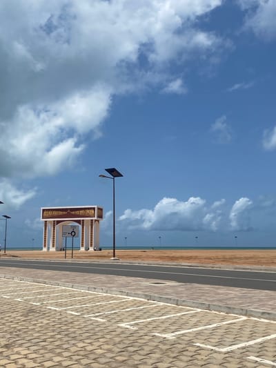 The image shows a partly cloudy sky above a paved parking area. A sign with the text "DJIGBADJI" and directions for "COTONOU VILLE" is visible. The edge of a beach can be seen in the background. The presence of street lamps suggests a public space. The location is Djègbadji, Benin.