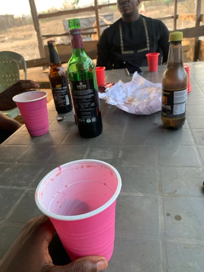 A table covered in various beverages. Bottles of wine and beer are scattered across the surface, alongside pink and red plastic cups. Some of the cups appear to have been used. A crumpled piece of white paper sits near the wine bottles. One person is wearing a dark shirt and facing away from the camera. Another person's arm is visible in the lower right corner, holding a pink cup. The setting appears to be outdoors, with trees and sky visible in the background.