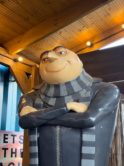A life-sized statue of the animated character Gru, from the "Despicable Me" franchise, is prominently displayed within a covered structure in Felixstowe, United Kingdom. The statue, depicted from the upper torso, features Gru with his arms crossed over his chest, a wide, closed-mouth smile, and his gaze directed slightly to the viewer's left. He wears his signature grey-striped scarf and a dark grey or black jacket with a visible vertical silver zipper. His head is bald, and his facial features include a large, pointed nose and light-colored eyes.
The immediate environment consists of a robust wooden canopy. The ceiling is constructed from exposed wooden planks and beams, stained a warm brown, with multiple bare incandescent light bulbs providing illumination. To the statue's left, a vertical wooden beam supports the structure, with a single light bulb attached. Behind this beam, a surface of light blue corrugated metal or paneling is partially visible, and below it, a portion of a sign with white text, reading "SETS THE," is discernible against a colorful background. To the right of the statue, a vertical screen or wall composed of numerous narrow, light-brown, bamboo-like slats or reeds extends upwards. The scene depicts a static, anthropomorphic subject within an artificially lit, sheltered setting.
