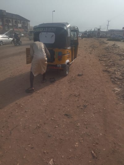 The image shows a man in a long, light-colored robe standing next to a yellow, black, and red auto rickshaw. The rickshaw's license plate reads "KKAR-00781." The man is pushing the rickshaw with his right hand, and his left hand is resting on the side. The rickshaw is parked on a dirt road with a few buildings in the background. The road is in poor condition, with loose rocks and gravel covering the surface. The scene appears to be in a residential or commercial area. The coordinates, latitude: 9.029, longitude: 7.584, correspond to a location within the city of Way, Nigeria.