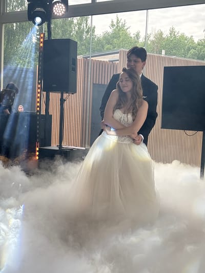 A man and a woman are embracing in front of a wooden wall with large windows. The woman is wearing a white wedding dress, and the man is wearing a dark suit. They are surrounded by low-lying fog or smoke, illuminated by stage lights. In the background, a DJ setup with speakers and lighting equipment is visible. The overall scene appears to be at an event, possibly a wedding reception. The location is identified as Osipovo, Russia.