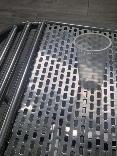 The image displays a close-up, overhead view of a transparent plastic cup filled with a dark liquid and ice, resting on a metallic grated surface. The cup is positioned to the right of the frame. The metallic surface has a pattern of oval-shaped perforations and appears to be part of a larger structure, possibly a table or a tray. To the left of the grated surface are several parallel, shiny metallic tubes, suggesting a supporting frame. The background is a dark, textured surface, possibly a floor or wall. There are no people visible in the image. The scene is indoors or in a covered outdoor area, given the lighting and the nature of the objects. The location of Sokuluk, Kyrgyzstan is not discernible from the visual elements present in the image.