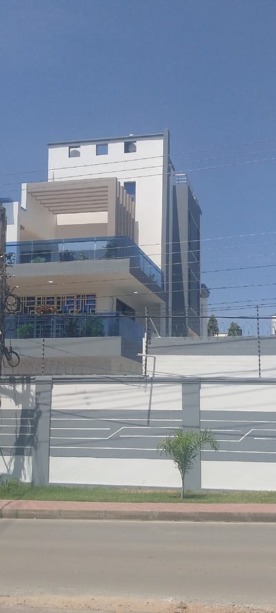 The image depicts a modern, multi-story building located in the Ziwa La Ng'ombe ward, Kenya, viewed from a street-level perspective on a clear day.
In the foreground, a gray asphalt road occupies the bottom portion, bordered by a reddish-brown paver curb. Behind the curb is a narrow strip of green grass, where a small, young palm tree is planted slightly off-center to the right.
Immediately behind the grassy strip stands a perimeter wall, predominantly white with horizontal gray patterns. The wall is topped with multiple strands of razor wire and an electric fence system, evidenced by white support posts and insulators. Shadows from utility lines are cast across the wall.
The central subject is a contemporary building featuring a white and gray facade. The lower visible floor has blue-tinted windows with white security grilles. Above this, a large balcony is present, enclosed by a blue-tinted glass railing, displaying several potted green plants. A prominent beige pergola-like structure with horizontal slats covers part of this balcony. Further up, the building exhibits various architectural elements, including vertical gray and white paneling on its right side, additional windows, and a flat roofline with small rectangular cutouts. No people are visible within or around the building.
Multiple black utility lines traverse horizontally across the mid-section of the frame, partially obscuring the building and the perimeter wall. A utility pole is partially visible on the far left. The sky above is clear and light blue.