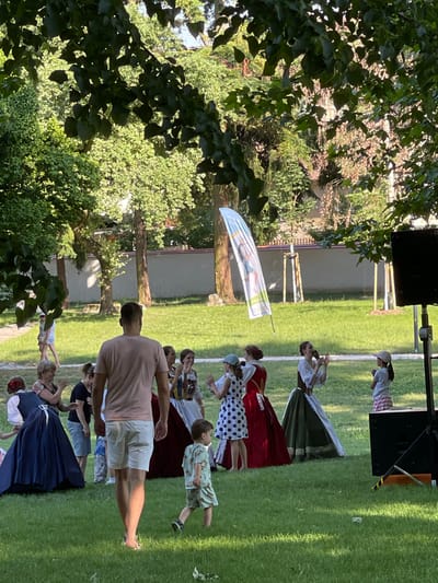 This is a photograph of an outdoor gathering, possibly a festival or performance, taking place in Stupava, Slovakia. The scene is set on a grassy area with trees and foliage providing a natural backdrop. A stage or platform with a speaker is visible in the upper portion of the image, suggesting a performance is underway. Several people are present, some in traditional or costume attire, including women in long skirts and dresses, and a child in patterned shorts. Others are dressed more casually in t-shirts and shorts, suggesting they are performers or attendees.
The activity appears to be a group of people, likely performers, engaged in a coordinated movement or dance. One individual in the foreground is a man in a light pink t-shirt and white shorts, his back to the camera. Several other individuals are scattered across the grass, some appearing to be watching or participating. A flag with some graphical elements and text is displayed, fluttering in the breeze. The lighting suggests it is daytime, likely late afternoon or early evening given the warm tones. The overall atmosphere seems lively and festive.