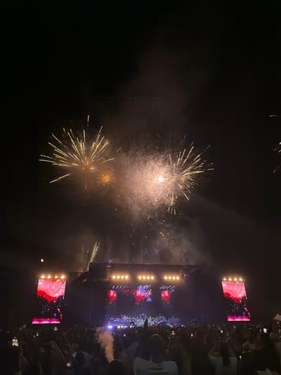 The image depicts an outdoor concert at night, set against a dark sky illuminated by fireworks. A large stage is visible in the background, featuring multiple screens displaying vibrant red and pink graphics, and a band performing on stage. A massive crowd of people stands in silhouette in the foreground, many holding up their phones to capture the event. The overall atmosphere is festive and energetic, with the fireworks adding a spectacular element to the concert. The location is the main square of Mosta, Malta, during the annual feast celebrations.