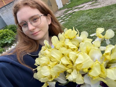 The image is a brightly lit outdoor portrait, likely a selfie, of a young woman holding a large bouquet of yellow irises. The photograph is oriented with the subject's head tilted towards the bottom left, appearing rotated 90 degrees clockwise from a standard portrait.
The main subject is a young woman with long, wavy reddish-brown hair and visible freckles across her face. She is wearing modern, thin-framed silver glasses and has a calm, neutral expression. She is dressed in a dark blue garment, possibly a sweatshirt or jacket, and a delicate silver chain is visible around her neck.
She is holding a dense bouquet of numerous yellow irises. The petals are predominantly a soft, pale yellow. Closer inspection reveals that some of the inner petals, particularly the 'falls' (lower petals), are lighter, almost white, with distinct brown or purple striped patterns.
The background suggests an outdoor setting, likely a garden or yard in Новая Тура, Russia. To the left (when viewed upright), there is lush green grass and a dirt or gravel path. A light-colored brick structure, possibly a wall or building, is partially visible. To the right (when viewed upright), there is green foliage from bushes or trees, and a grey, textured surface that appears to be a roof or another building element. The bright, even lighting indicates it is daytime, likely a clear day. No visible text is present in the image.