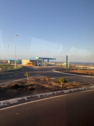 An outdoor, eye-level shot captures a gas station and car wash facility under a clear blue sky in Puerto del Rosario, Spain. The facility features a blue and green covered canopy with "moeve" branding. An orange van is parked near the pumps, and a dark-colored car is also visible. The surrounding landscape includes a paved road, a white-lined curb, and sparse, dry vegetation. In the background, the ocean stretches to the horizon, with several lamp posts and other structures suggesting an urban or industrial area near the coast. The image appears to have been taken from a moving vehicle, indicated by reflections on the surface.