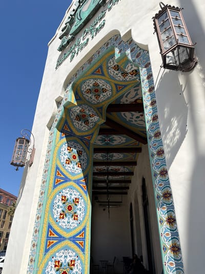 The image shows the facade of a building with ornate tilework around an arched entryway. The tiles are a vibrant mosaic of blue, yellow, red, and white geometric and floral patterns. Above the archway, a sign with raised, green lettering is partially visible against a clear blue sky. Two wrought-iron lanterns with glass panels are attached to the white stucco wall, one on the left and one on the right of the entryway. The lantern on the right casts a shadow of intricate latticework onto the wall. Inside the entryway, wooden beams are visible on the ceiling, also decorated with colorful tiles. In the background, through the archway, a dimly lit interior with tables and chairs can be seen, and a person is seated inside. To the left of the building, a few floors of an adjacent building with balconies and a palm tree are visible. The overall scene suggests a sunny day in Pasadena, California, likely a historic district known for its Spanish Colonial Revival architecture, as exemplified by the elaborate tiling and architectural details.
