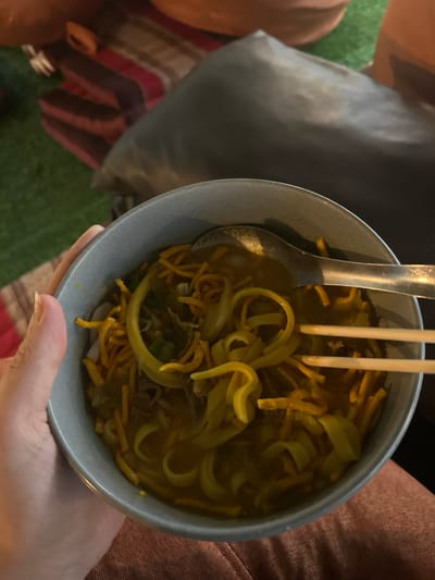 The scene is set in Ban Paem Bok, Thailand, depicting a close-up, top-down view of a light grey ceramic bowl of food held by a person. The bowl contains a yellow-orange broth-based noodle dish, characteristic of Khao Soi, with both wide, flat egg noodles and crispy fried egg noodles visible, along with green garnishes like chopped scallions. A silver metal spoon is partially submerged in the broth on the left side of the bowl. A pair of light-colored wooden chopsticks are positioned upright within the noodles on the right side, suggesting active consumption or preparation to eat. A human left hand, with fair skin and visible fingernails, holds the bowl from the bottom-left edge. A portion of a leg, covered in reddish-brown fabric, is visible in the lower-right corner, belonging to the same individual. The background, dimly lit, features a dark grey cushion or pillow behind the bowl, partially revealing a striped textile (red, brown, and dark tones) and a green surface, possibly artificial turf or a mat, in the upper-left quadrant.