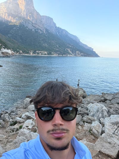 The image is a selfie taken by a young man with dark, wavy hair and a goatee, wearing sunglasses and a blue collared shirt. He is standing on a rocky coastline with the sea and a large, vegetated mountain range in the background. The sky is clear and blue. In the distance, two figures can be seen standing on rocks by the water. The scene is located in the area of Baunei, Italy, known for its dramatic coastal cliffs and turquoise waters.