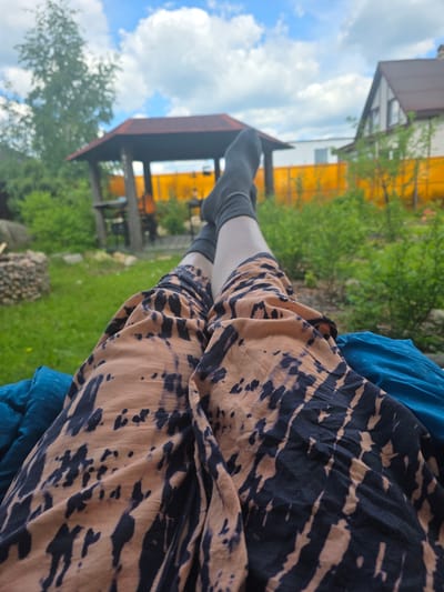 In a suburban outdoor setting, a person is lying down, with their legs extended towards the viewer. They are wearing black socks and patterned pants with a tie-dye design in brown and black. The person is resting on a blue surface, possibly a blanket or cushion. In the background, a wooden gazebo with a red roof is visible, along with green bushes and trees. Further in the background, a house with a brown roof and white siding, and a bright orange fence are present. The sky is blue with fluffy white clouds. This scene appears to be in Brilliant, Russia.
