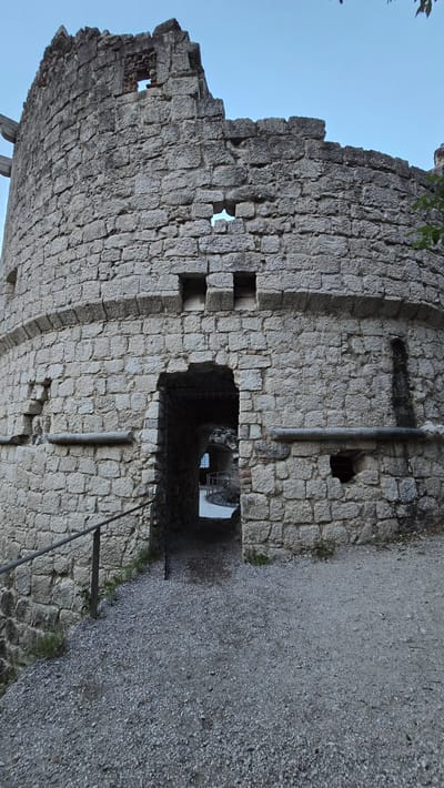 This is a photograph of a weathered stone structure, likely a castle or fortress ruin, against a clear blue sky. The stone is rough-hewn and irregular, with visible mortar between the blocks. The structure has a curved wall on the left and a more vertical section on the right, with several openings or windows visible. One large arched opening leads into a dark interior passage, where the faint outline of stairs and a railing can be seen. A gravel path runs along the right side of the structure, bordered by some sparse vegetation. The lighting suggests it is daytime, possibly late afternoon or early morning, given the long shadows. The overall impression is one of age and abandonment, with a sense of history and mystery. There are no people visible in the image. The location is Riva del Garda, Italy.