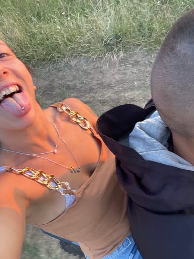 In an outdoor setting with dry, light green grass in the background, a young woman is shown from the chest up, taking a selfie. She has her tongue out, revealing a tongue piercing. She is wearing a tan, ribbed tank top with decorative gold chain straps. A silver necklace with a small diamond pendant and another chain with a word pendant hang around her neck. The image is cropped to include the back of a man's head on the right side of the frame. The lighting suggests it is either sunrise or sunset, casting a warm, orange glow on the woman. The location is stated to be Zheglartsi, Bulgaria.