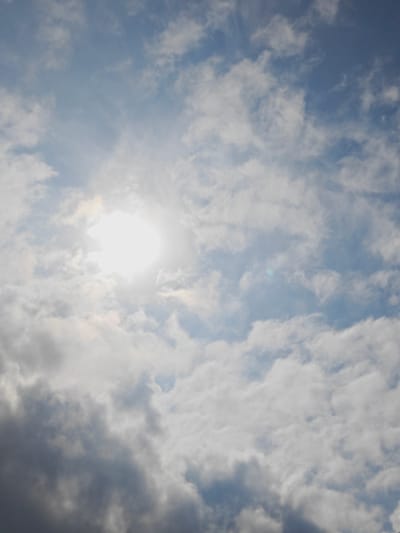 The image shows a bright, overcast sky with the sun partially obscured by clouds. The sky is a mix of light blue and white, with fluffy, scattered clouds. The sun appears as a bright, white disc in the upper left quadrant of the image, with rays of light radiating outwards. The lower portion of the image features darker, more dense clouds. There are no discernible buildings, people, or other objects visible in the image, indicating it is a photograph taken outdoors, focusing solely on the sky. The location of Koledino, Russia is not evident from the visual content of the image.