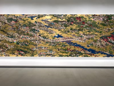 This image captures a section of the artwork "View of the Sea of the Genkai" by the artist Tōitsu. The artwork is displayed in a museum setting, likely at the Chichu Art Museum on Naoshima Island, Japan, given the characteristic minimalist white walls and display platforms visible. The artwork itself is a large, multi-panel screen rendered on a gold leaf background. It depicts a densely populated, vibrant urban scene, possibly a festival or bustling marketplace, with numerous small figures engaged in various activities. Buildings with red roofs and tiled eaves are interspersed with lush green trees and golden cloud-like motifs. A prominent blue river or body of water winds through the composition, crossed by several bridges. The artwork is illuminated by soft, overhead lighting, and the overall impression is one of intricate detail and a lively, energetic atmosphere.
