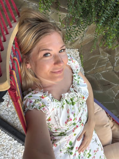 A Caucasian woman with blonde hair pulled into a bun, wearing a white dress with a floral print, lies in a hammock. She is looking at the camera and smiling. The hammock is made of colorful fabric and is suspended by wooden beams. In the background, there is a stone wall and green foliage. The scene appears to be outdoors, possibly in a garden or patio area. The location is specified as Nizhnyaya Shilovka, Russia.